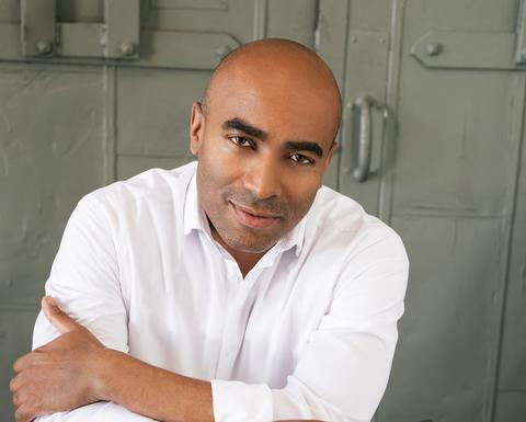Lee Hawkins dressed in sitting backwards on a chair and looking at the photographer with glasses in hand
